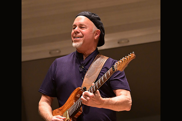 A smiling man wearing a dark cap plays an electric guitar.