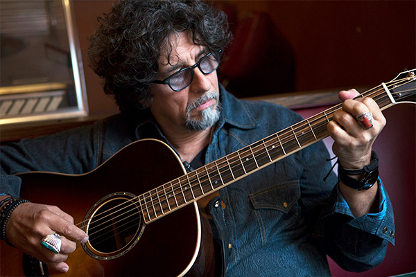A seated man in a denim shirt and glasses playing a guitar.