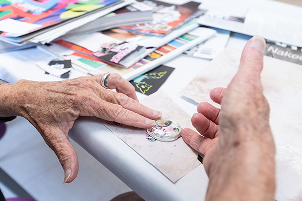Hands creating a small artwork at a table, with stacks of paper and magazines nearby.