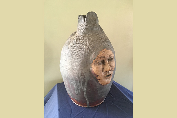 Vase-like ceramic vessel with a human face in one side, sitting atop a blue cloth.