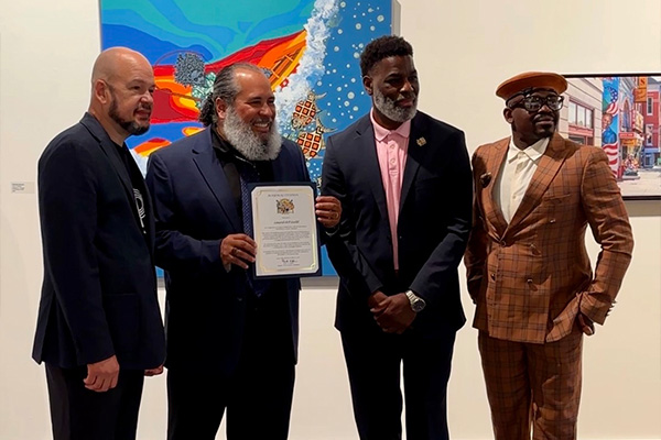 Four men in suits pose in an art gallery near a wall where colorful artworks hang. One of the men holds a certificate.