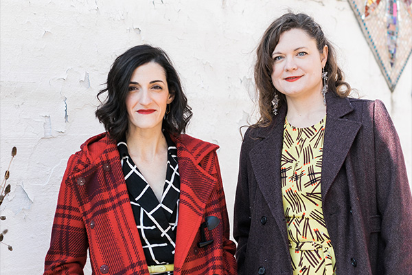 Two smiling women wearing patterned dresses and woolen overcoats.