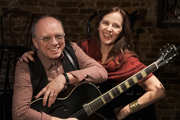A smiling woman in red poses with a smiling man in a black vest who is holding a black guitar.