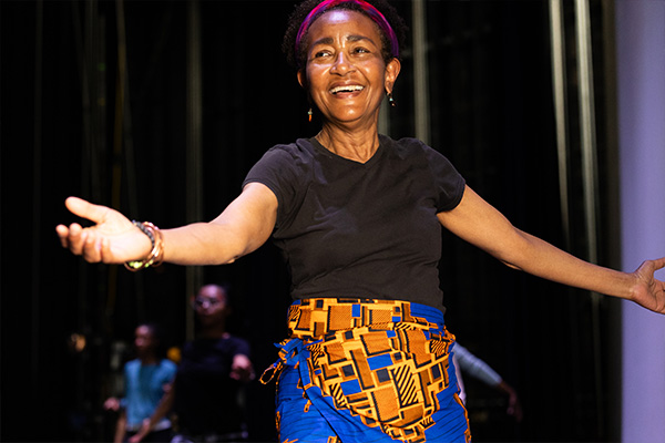 A smiling woman wearing a colorful wrap skirt stands on a stage with her arms outstretched at her sides.