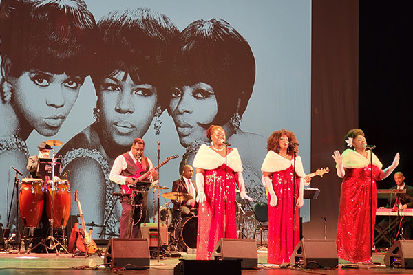 Three women in red gowns and white fur wraps sing into microphones, backed by a live band. A large photo of the Supremes is projected onto the backdrop behind them.