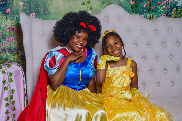 A child wearing a yellow satin princess costume poses next to a woman dressed as Snow White.