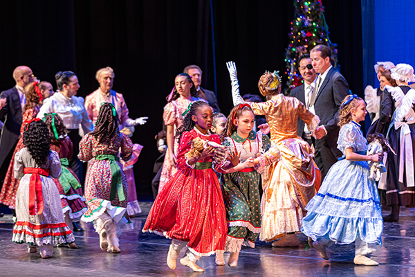 Costumed young performers in long dresses run across a stage near a decorated Christmas tree.