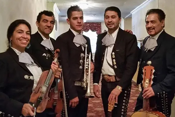 Five musicians wearing traditional black Mariachi suits with silver decorations pose holding their instruments, including a trumpet, violins, and guitars.