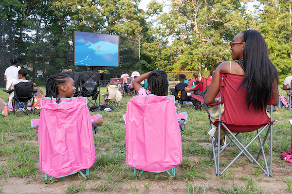 People sit in folding chairs outdoors, watching a movie on a large screen in a grassy park surrounded by trees.