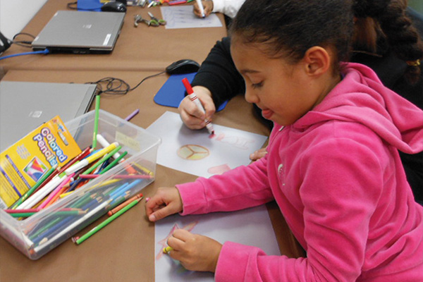 At a table, two people draw designs with markers and colored pencils on sheets of opaque Shrinky Dink plastic.