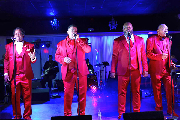 Four men wearing matching red suits sing into microphones on a stage.
