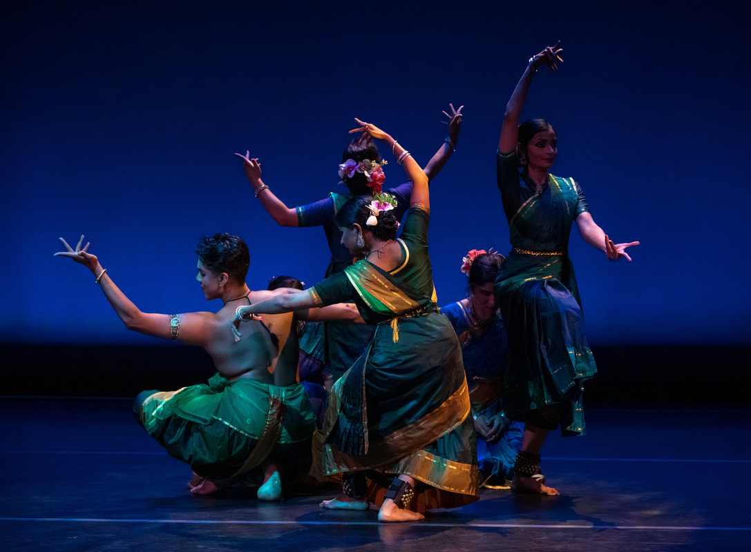 Group of dancers in green costumes perform a synchronized traditional dance on a stage illuminated by blue theatrical lighting.