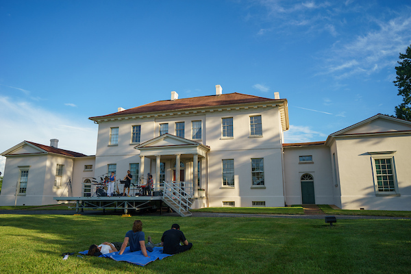 Outdoor concert at Riversdale House Museum, with attendees seated on a lawn facing a stage beside the historic mansion.