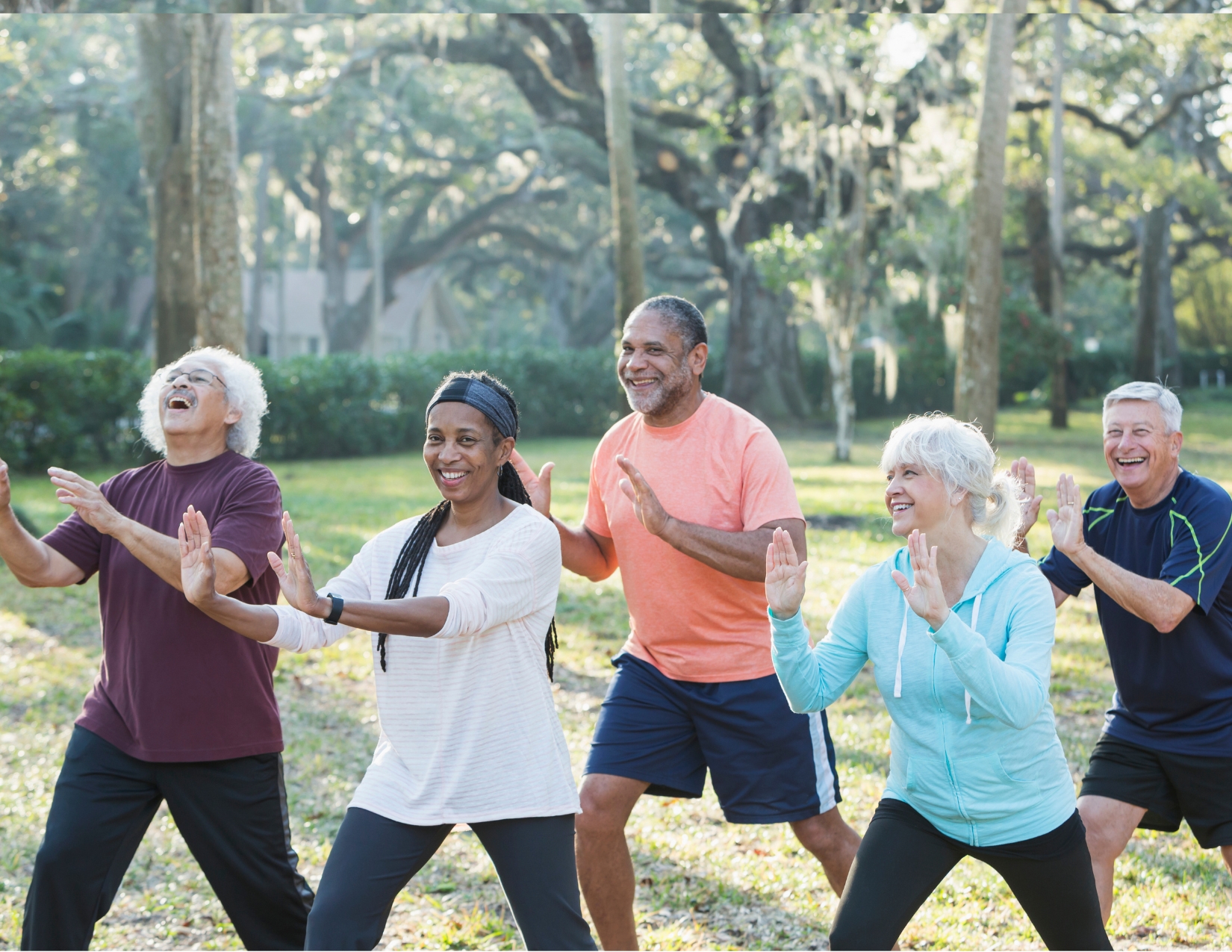 A diverse group of five smiling older adults practicing Tai Chi poses together in a sunny park surrounded by large trees.