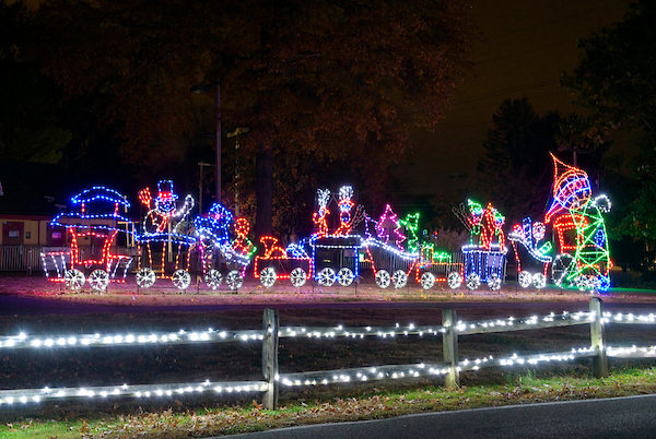 A festive holiday light display depicting a train loaded with toys and holiday items set up near a road at night.