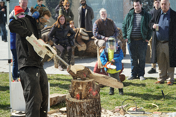 A person in protective gear carves away at wood using a chainsaw. People are gathered around to watch them work.